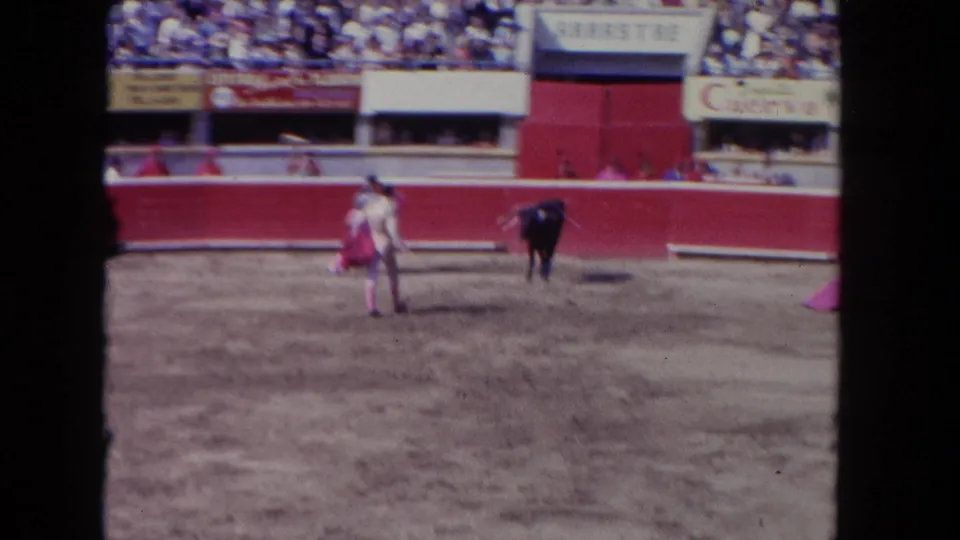 1962 Los Angeles 8mm frame showing a matador with a pink cape near a black bull as spectators watch from the stands.
