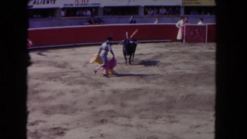1962 Los Angeles 8mm frame of a matador moving close to a black bull with capes visible in a red-walled arena.