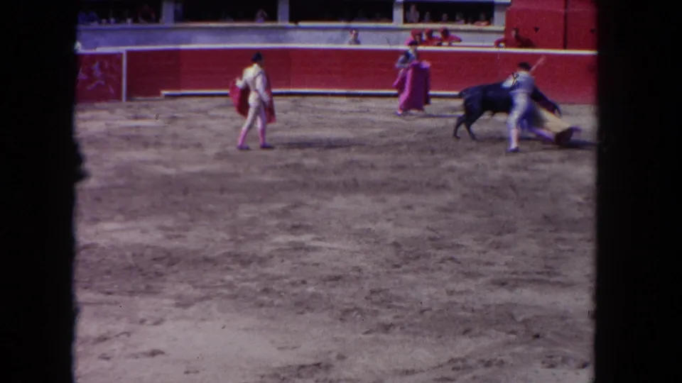 1962 Los Angeles home-movie frame showing matadors with capes facing a dark bull in a red-walled arena.