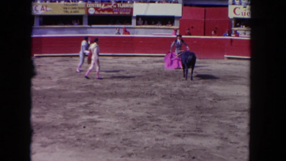 1962 Los Angeles bullring view with matadors, a dark bull, pink cape, spectators, and Spanish-language signs above the wall.