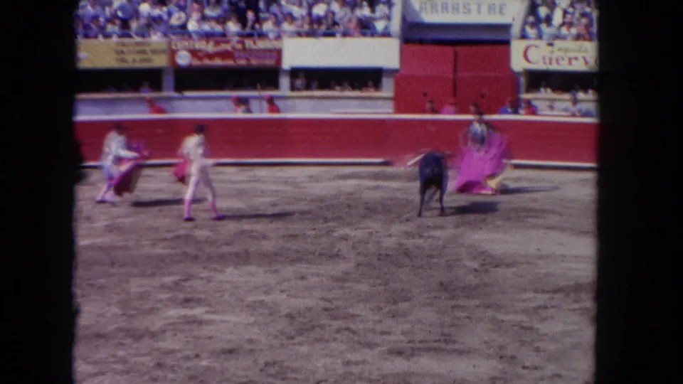 1962 Los Angeles arena frame showing matadors using capes around a dark bull, with advertising signs and spectators in the background.