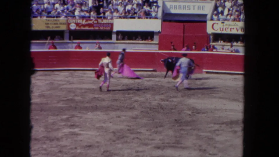 1962 Los Angeles home-movie frame with matadors moving across a sandy bullring as a dark bull stands near the red wall.