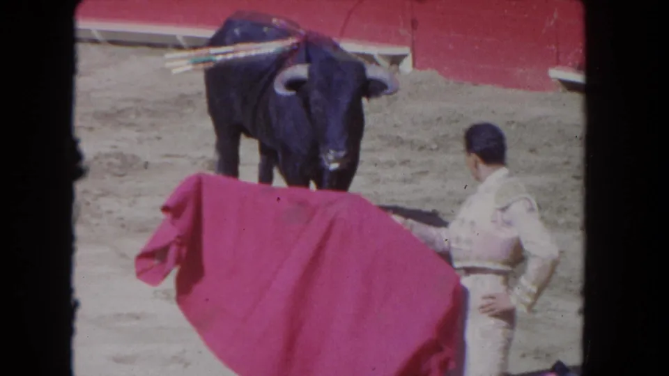 A matador in a light suit presents a red cape while a dark bull stands ahead in the arena.