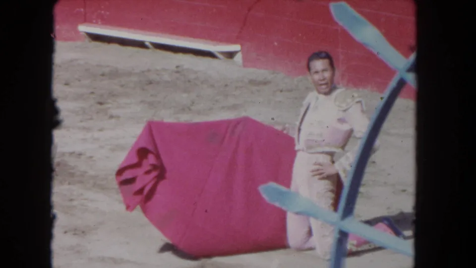 1962 Los Angeles 8mm frame showing a matador kneeling beside a red cape, with a red arena wall and blue railing visible.