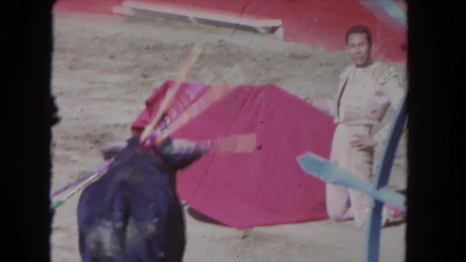 1962 Los Angeles 8mm frame with the bull in the foreground near a red cape and a kneeling matador behind blue barrier bars.