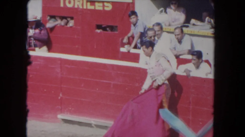 1962 Los Angeles 8mm frame showing a matador in a pale suit holding a red cape beside a red arena wall and spectators.