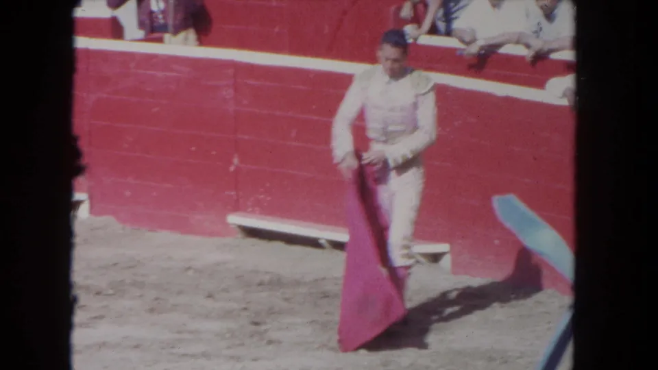 1962 Los Angeles frame of a matador holding a red cape on the sandy floor of a red-walled bullring.