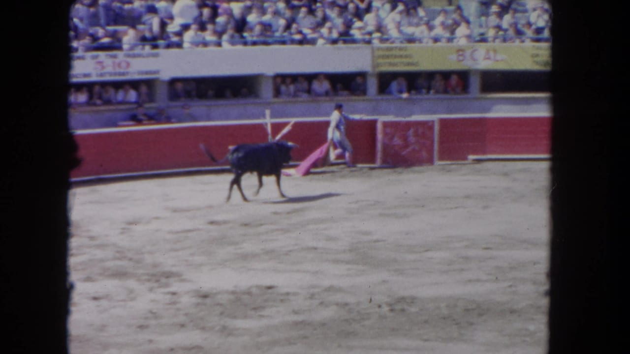 1962 Los Angeles 8mm frame showing a black bull crossing a sandy ring while a matador stands near the red barrier with spectators above.