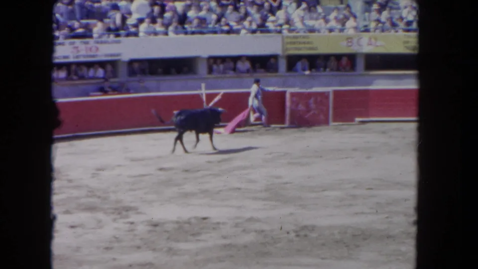 1962 Los Angeles 8mm frame showing a black bull crossing a sandy ring while a matador stands near the red barrier with spectators above.
