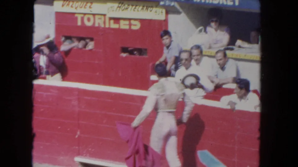 1962 Los Angeles 8mm frame showing a matador with cape near the arena wall, spectators close by, and a red TORILES door behind them.
