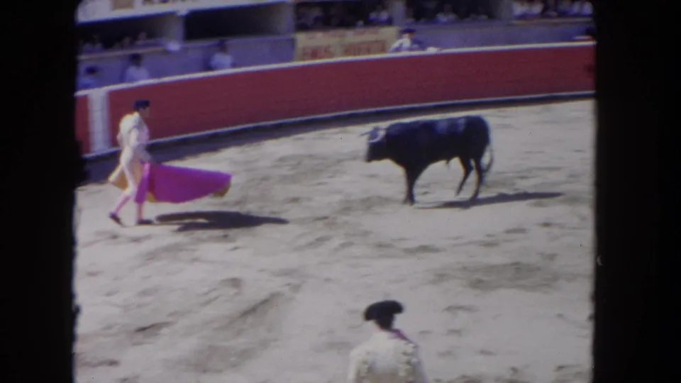 1962 Los Angeles home-movie frame showing a matador in white holding a pink cape toward a dark bull in a bullfighting arena.