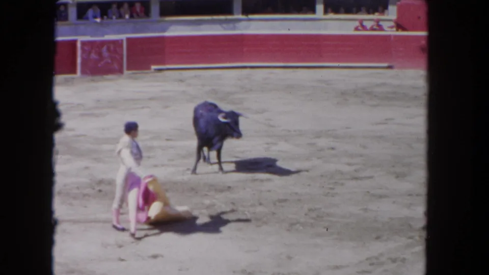 The matador lowers the bright cape as the black bull faces him in a sandy Los Angeles arena, recorded on 8mm film.