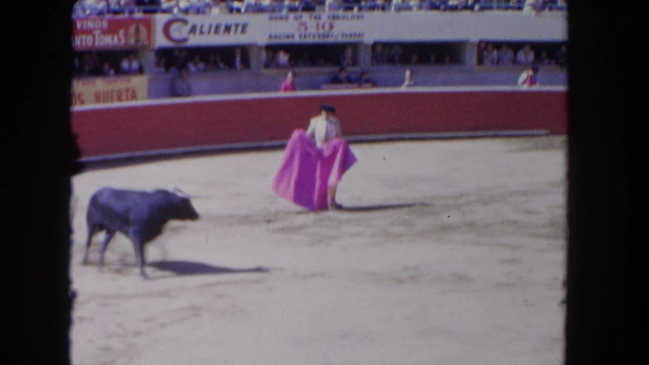 1962 Los Angeles home-movie frame showing a matador with a pink cape facing a black bull in a sandy bullring.