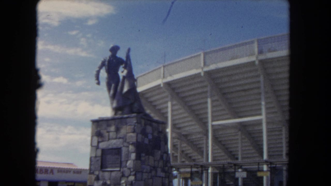 Matador statue on a stone base beside a stadium structure under a bright sky.