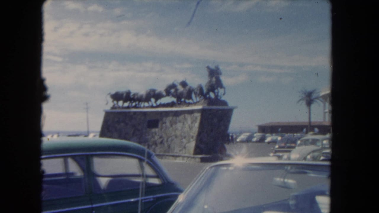 Stone monument with horse sculpture beside a parking lot filled with classic cars.