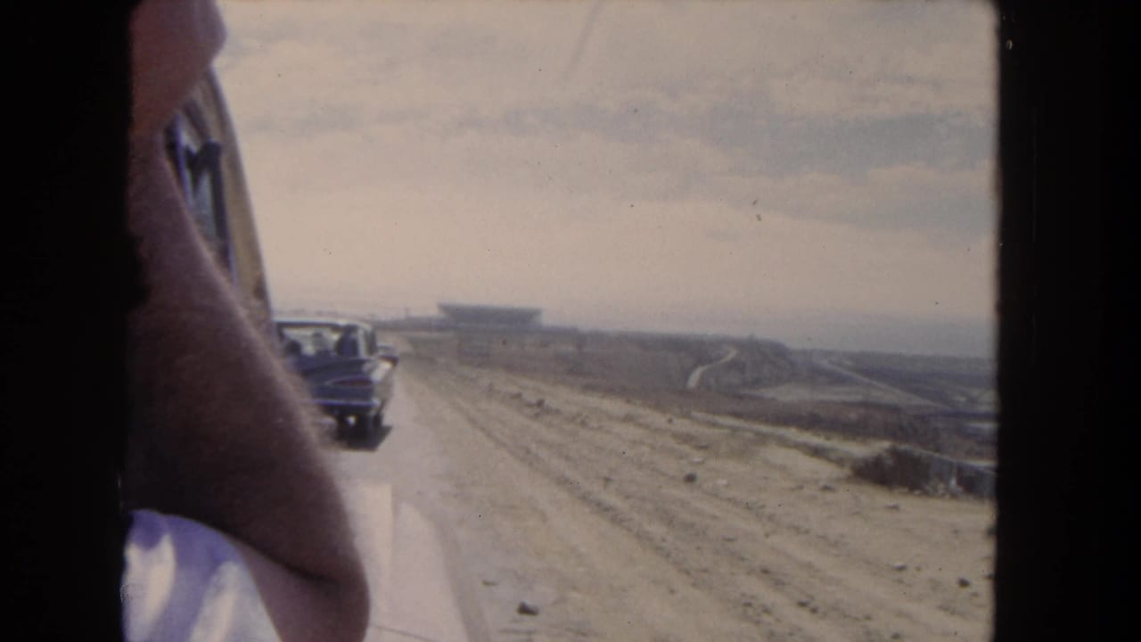 1962 Los Angeles home-movie frame looking past a passenger from inside a vehicle toward a car on a dirt road and open terrain.