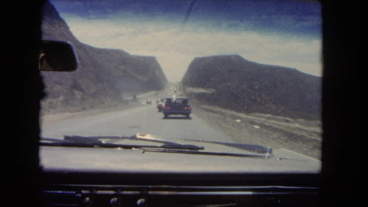 View through a car windshield of a highway between dry hills with cars ahead.