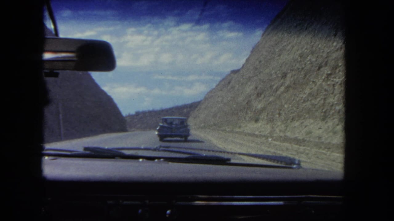Dashboard view of a car following a sedan on a paved road between rocky cuts.