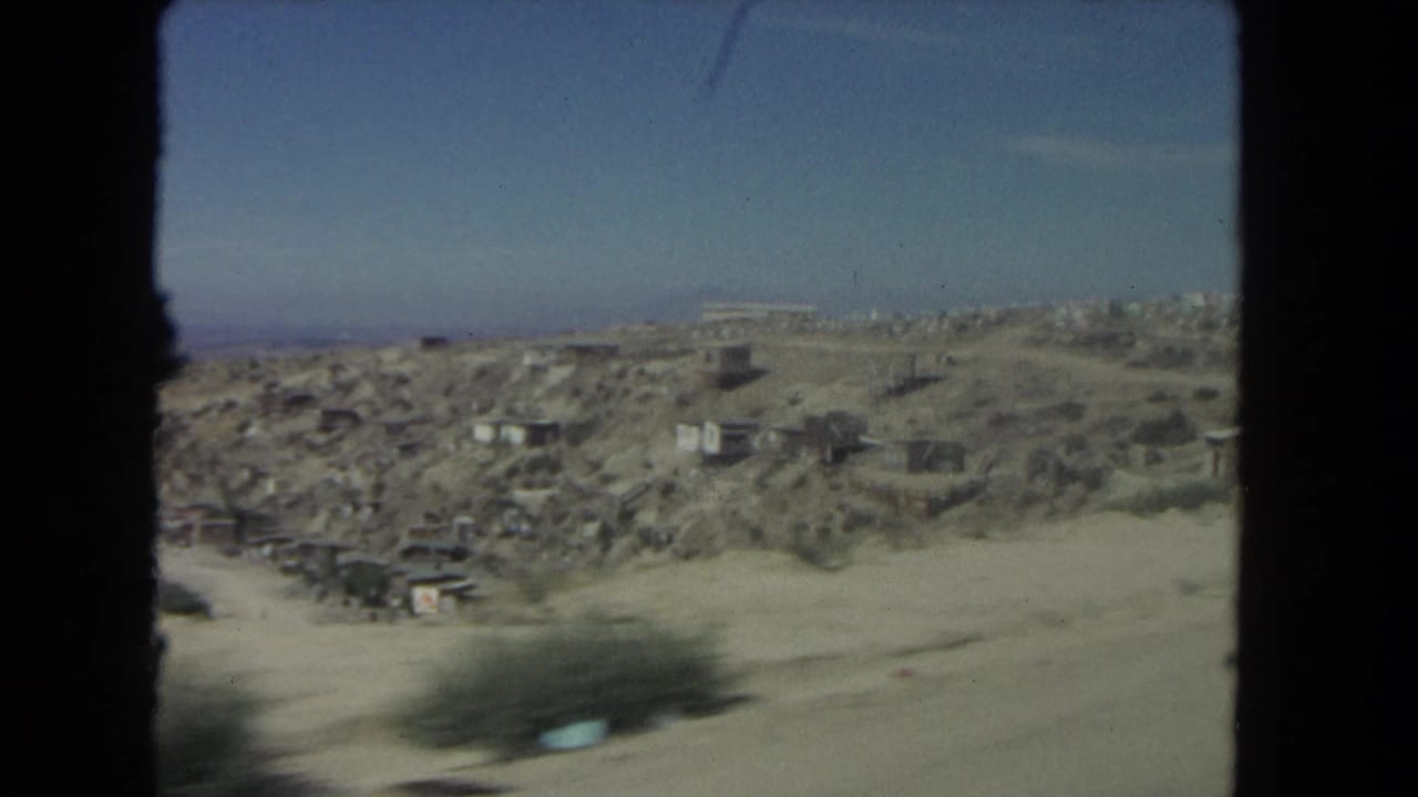 Dusty road beside arid hillside with scattered small structures under blue sky.