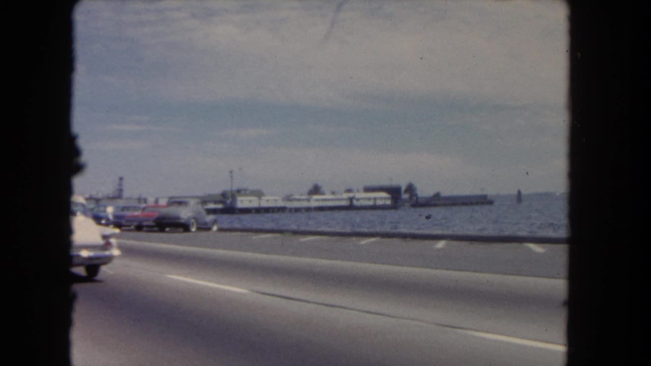 Cars travel along a waterfront road with a distant pier under a cloudy sky.