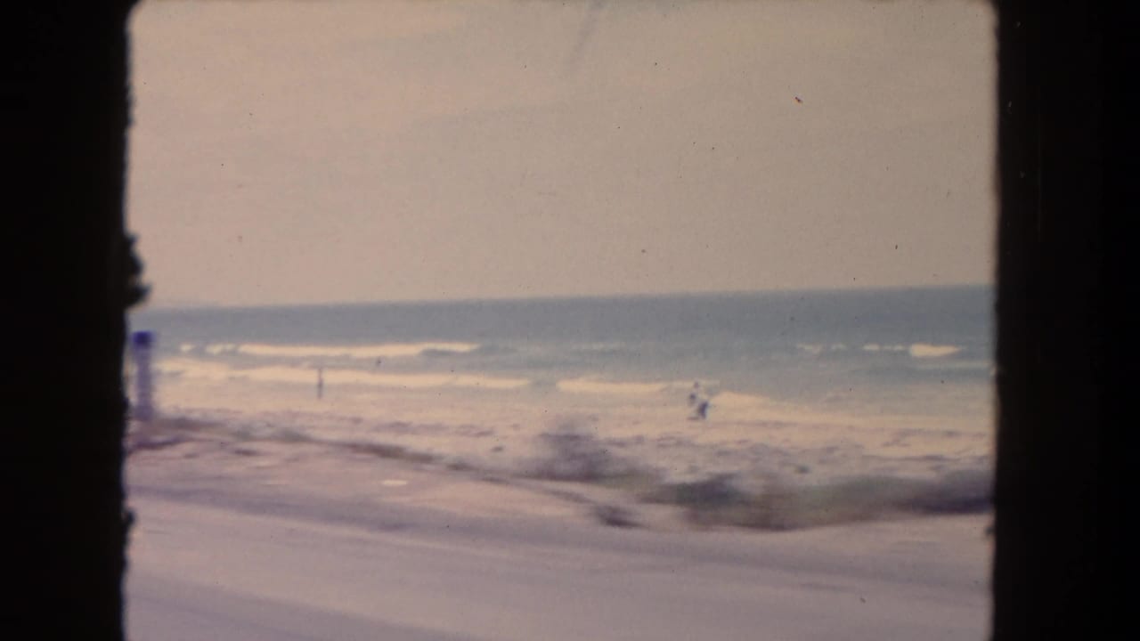 Surf and sandy shoreline with distant people in a soft 8mm frame.