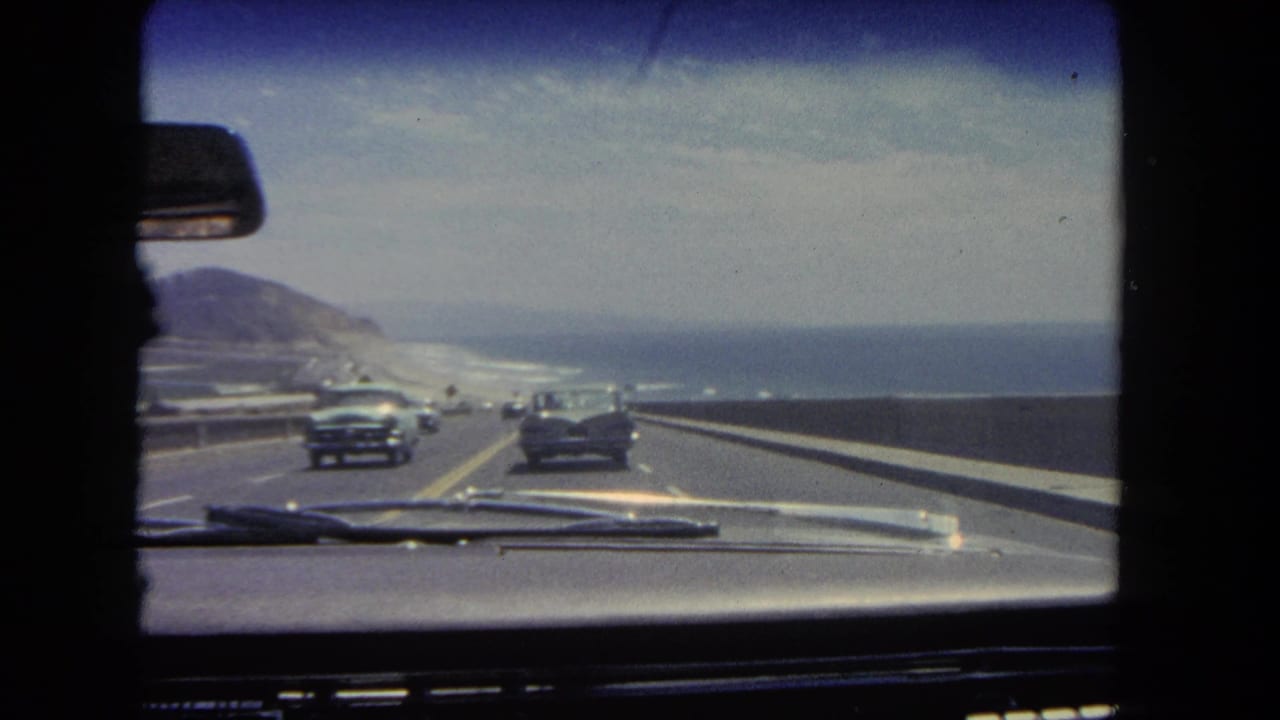 Coastal highway seen through a car windshield with classic cars and ocean beyond.