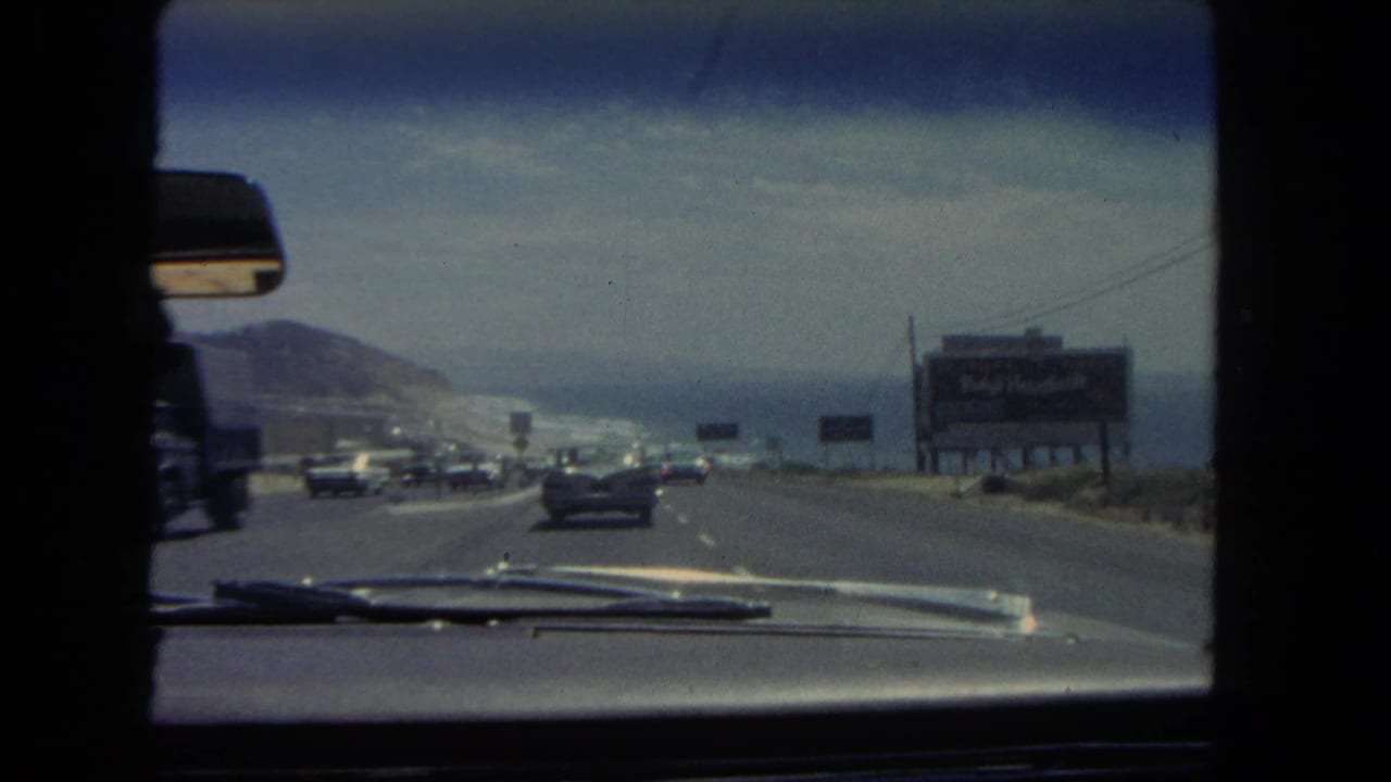 Windshield view of a coastal highway with cars, billboards, a hillside, and ocean beyond the road.