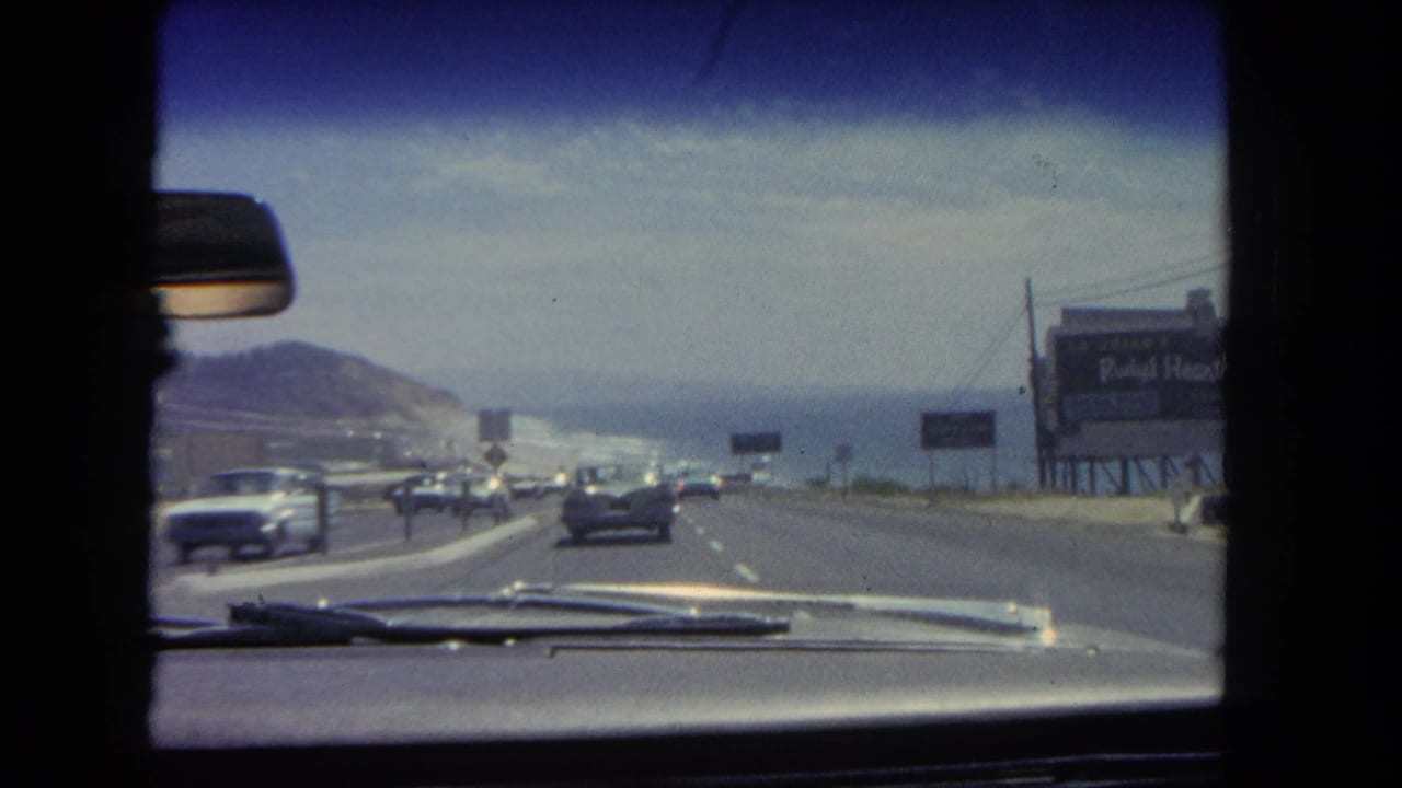 Car interior viewpoint looking down a divided coastal road with traffic, signs, hillside, and ocean.