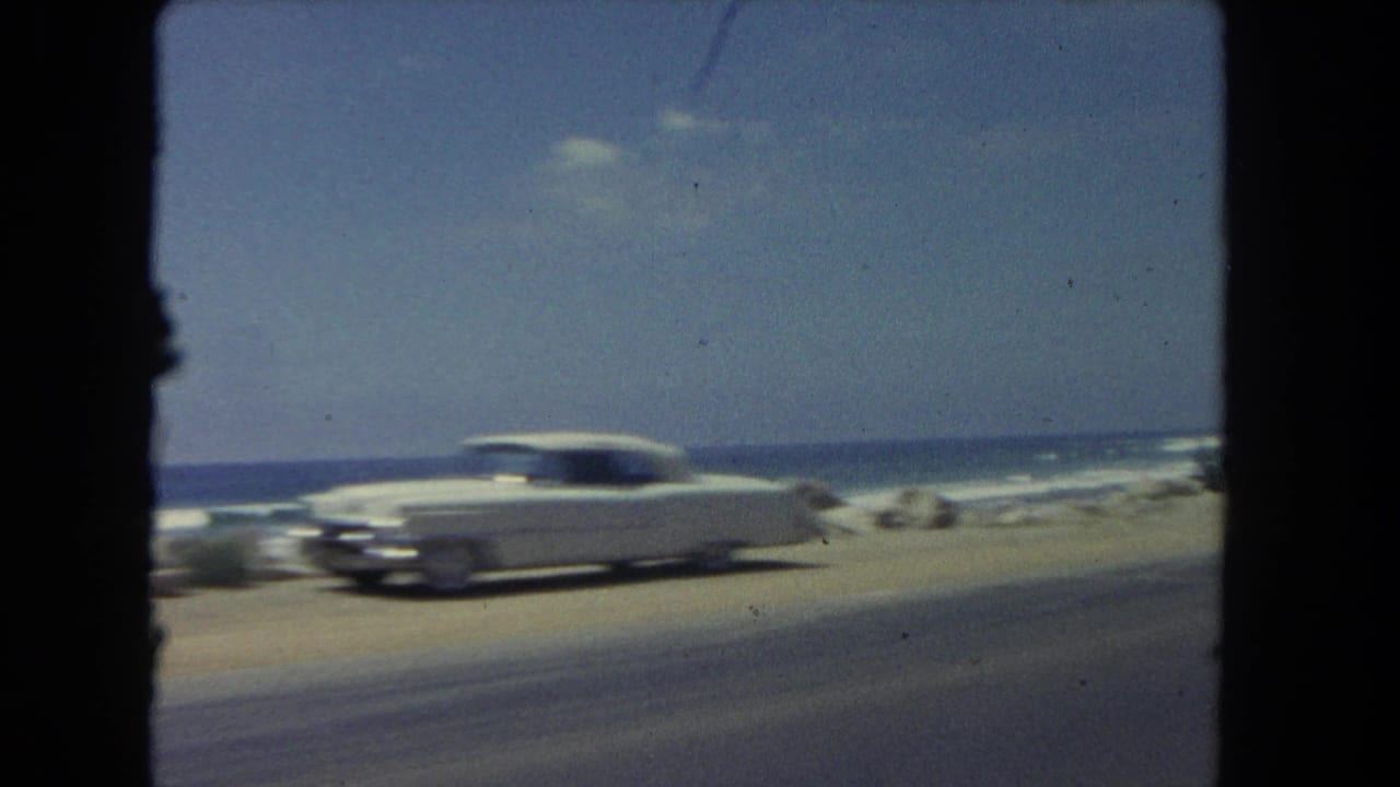 A white early-1960s sedan travels along a Los Angeles beachside road, with surf, rocks, and blue sky in the background.