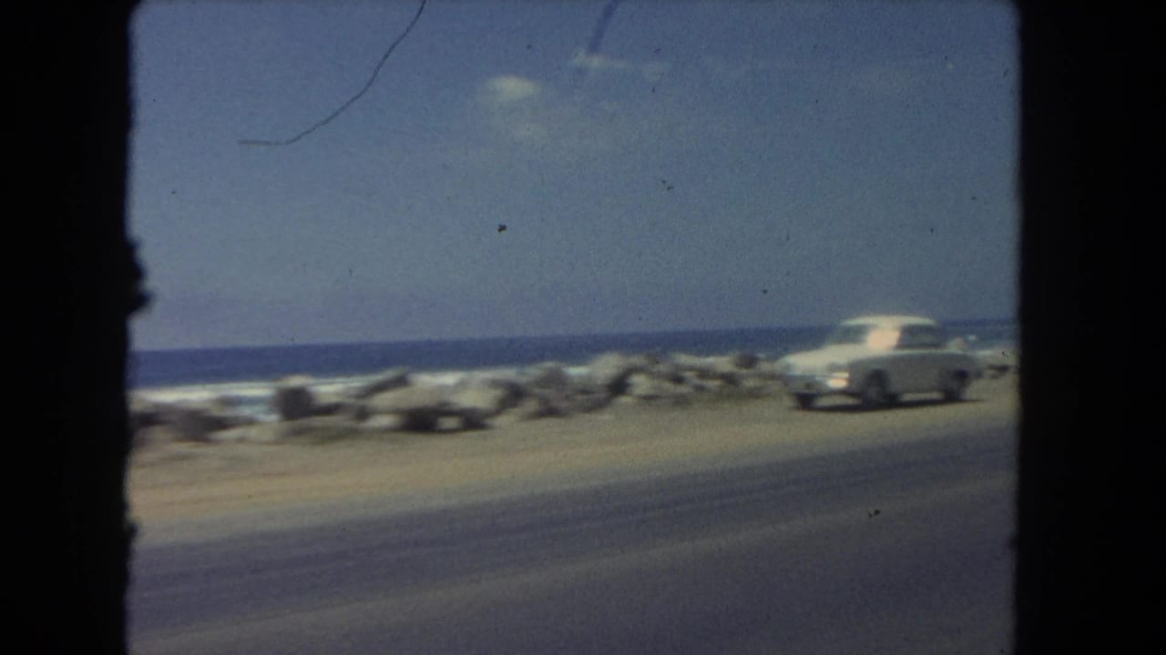 Beachside roadway with a white car near rocks and ocean surf.