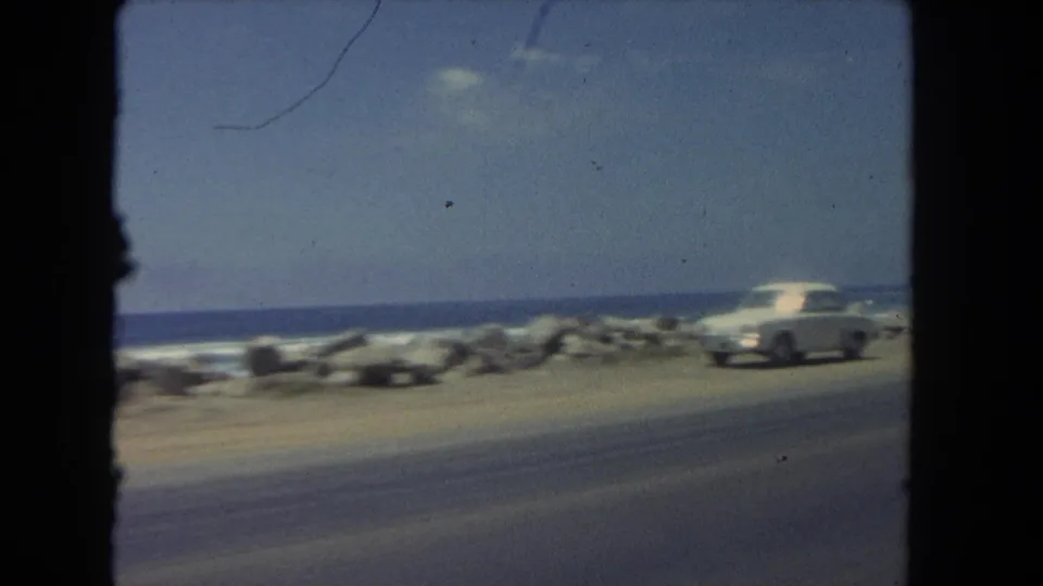 A white classic car appears near the edge of a coastal road in Los Angeles, with rocks, waves, and open sky behind it.
