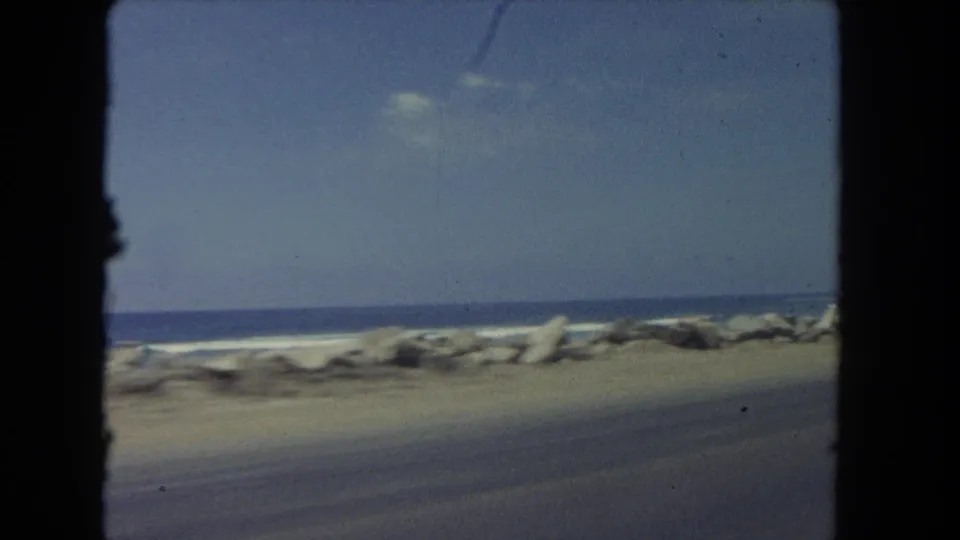 A moving-vehicle view along a Los Angeles coastal road shows the rocky breakwater, surf, sandy edge, and clear sky.