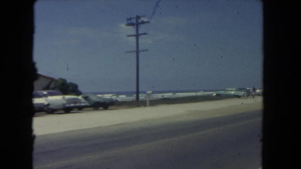 1962 Los Angeles coastal road view from a moving car, with parked cars, surf beyond the roadside, and a utility pole near the beach.