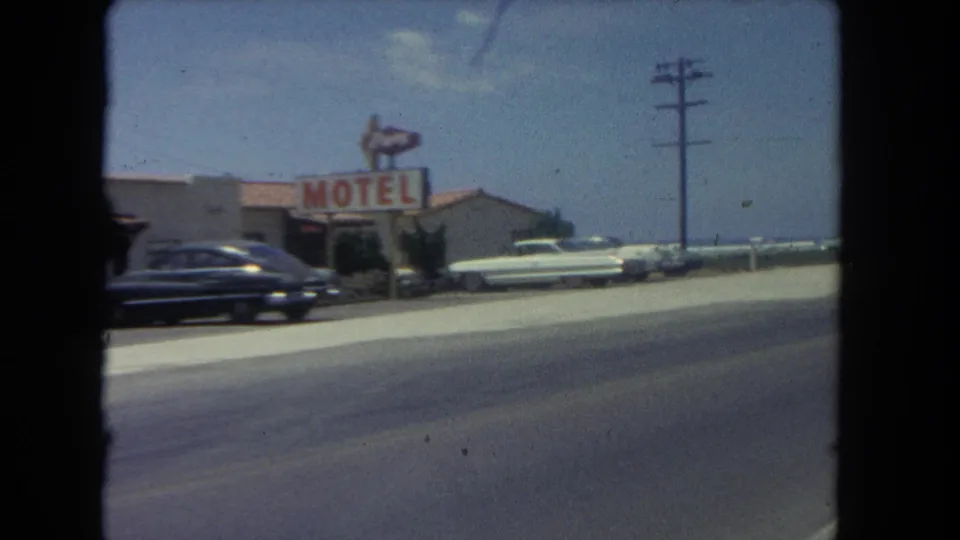 A 1962 Los Angeles roadside motel passes the camera, with mid-century cars parked out front and the ocean visible beyond the road.