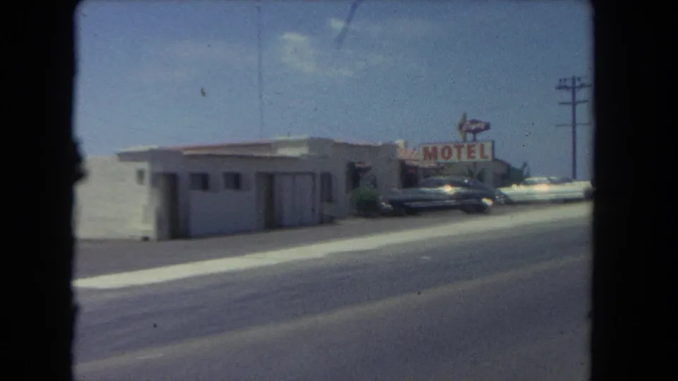 The moving view shows a low roadside building, a motel sign, parked cars, and a utility pole along the Los Angeles coast in 1962.