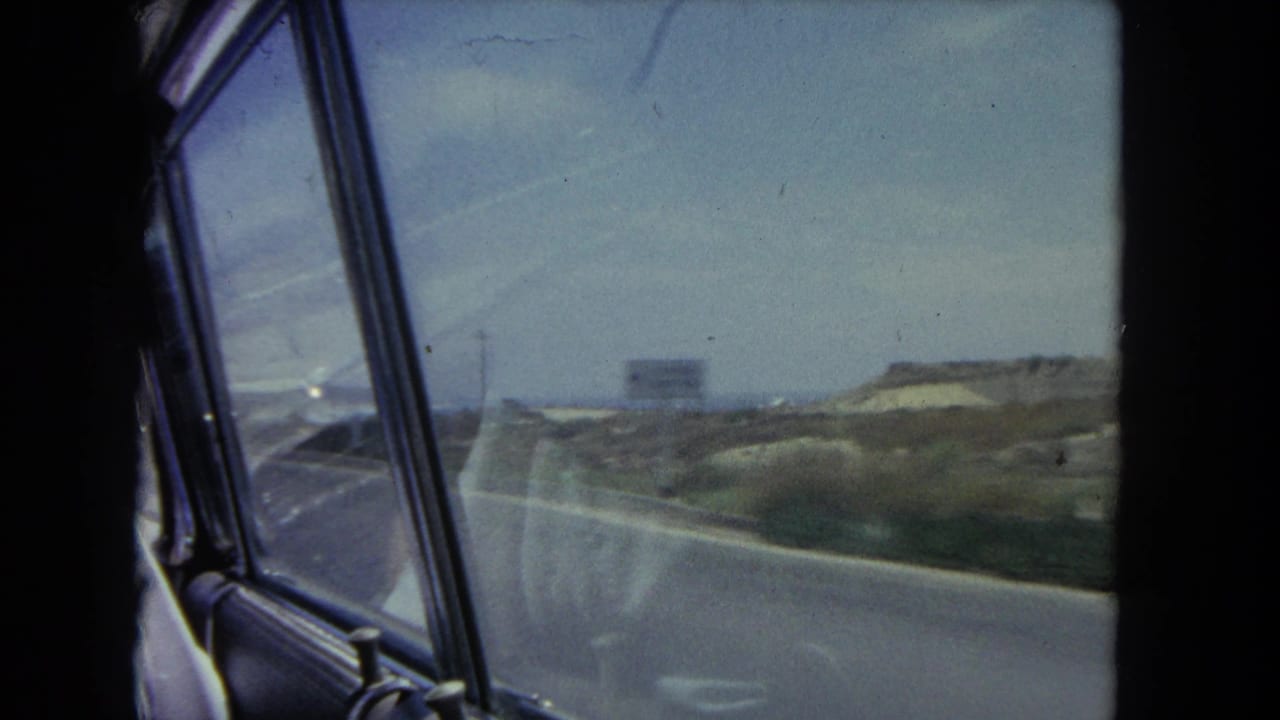 Passenger-window view of a coastal road with dunes, sky, and a glimpse of ocean beyond.