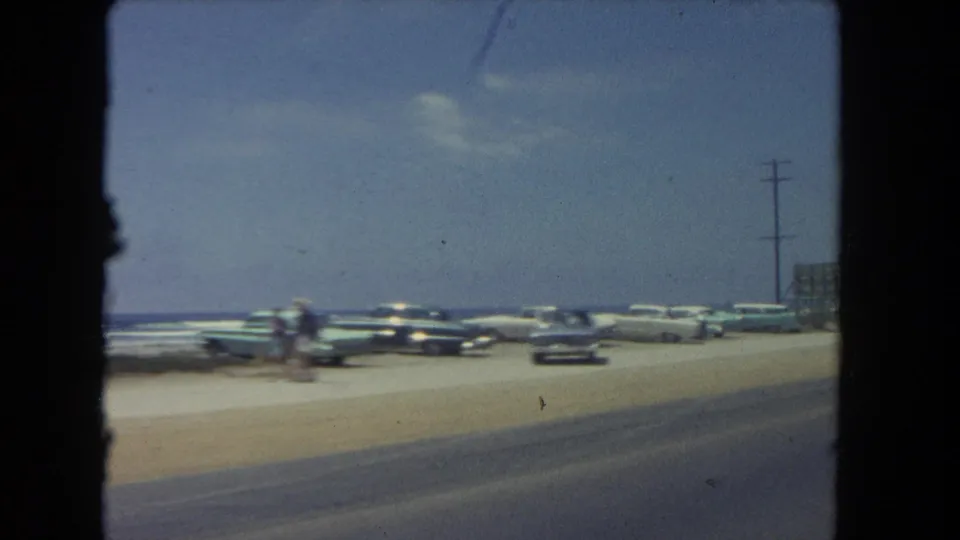 1962 Los Angeles beachside road scene with parked classic cars, small visible beachgoers, surf, and a roadside utility pole.