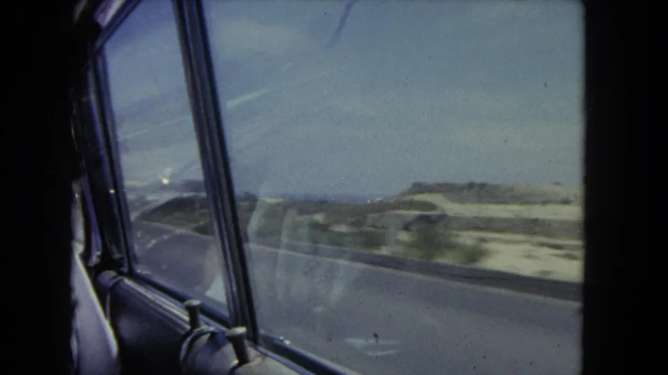 Moving car window frames a blurred view of coastal dunes and roadway in 1962 Southern California footage.