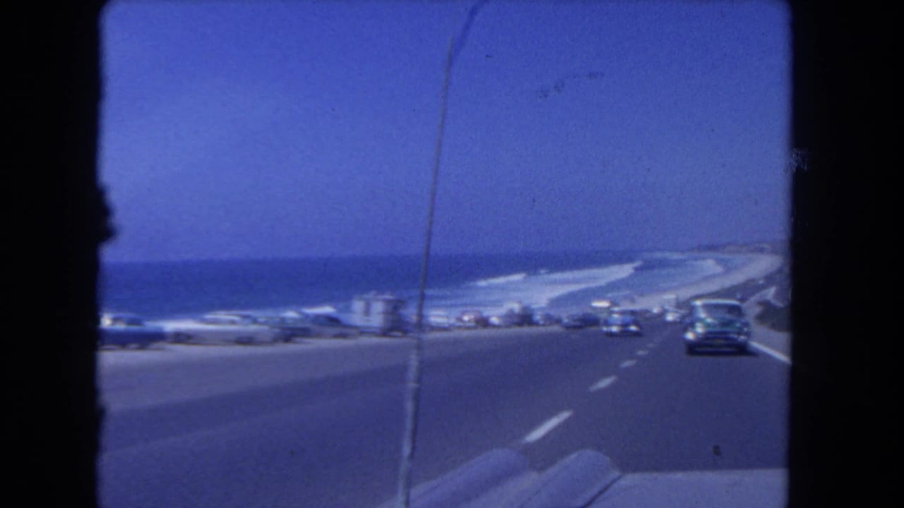1962 Los Angeles coastal highway view from a moving car, with classic traffic, roadside vehicles, blue sky, and ocean visible ahead.