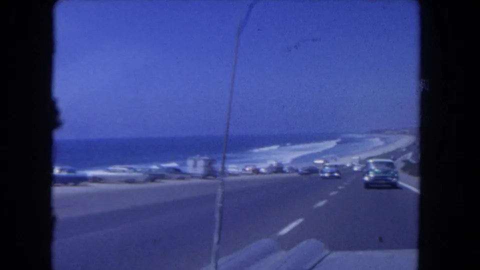 1962 Los Angeles coastal highway view from a moving car, with classic traffic, roadside vehicles, blue sky, and ocean visible ahead.