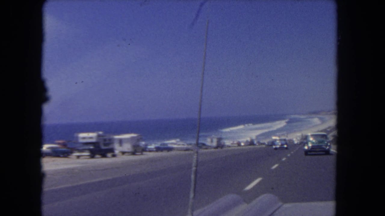 1962 Los Angeles coastal road scene showing parked RVs and cars near the ocean, viewed from inside a moving vehicle.