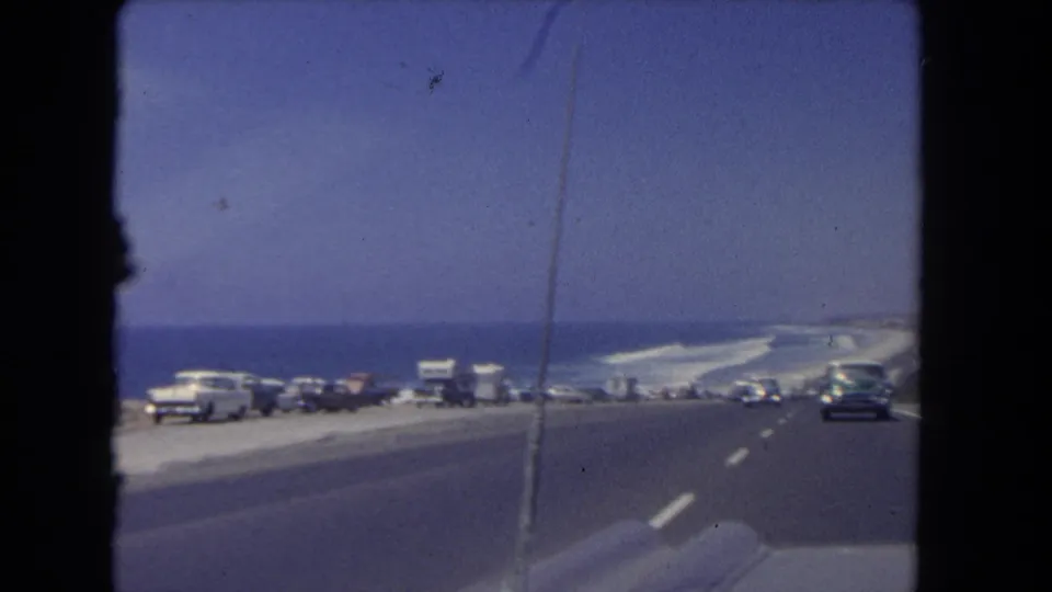 Los Angeles coastal highway footage from 1962, with a row of parked cars and RVs beside blue water and a broad road surface.