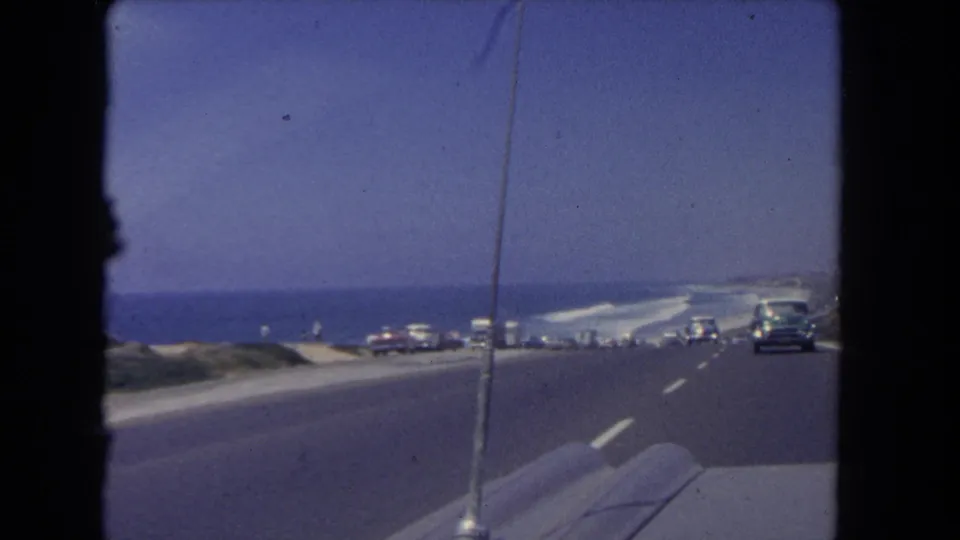 End frame of a 1962 Los Angeles coastal drive, showing ocean, roadway, distant parked vehicles, and a car ahead through the windshield.