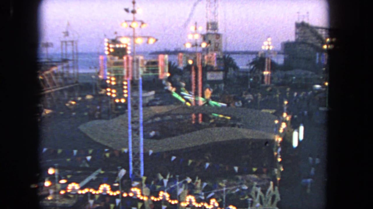 Wide night view of illuminated amusement rides, pennant flags, and distant visitors.