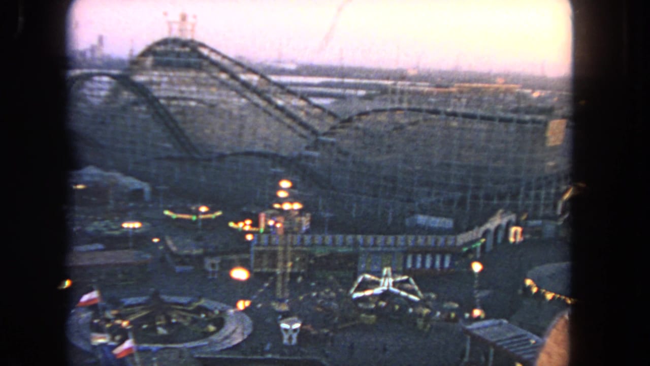 Dusk view over an amusement park with a wooden roller coaster and illuminated rides below.
