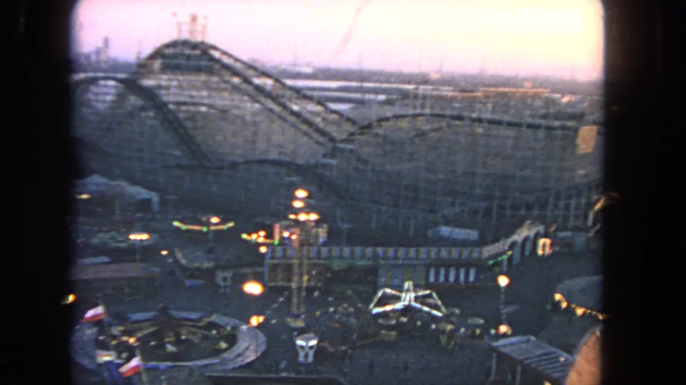 1962 Los Angeles 8mm view over an amusement park at dusk, with a large wooden roller coaster, midway lights, and visitors below.