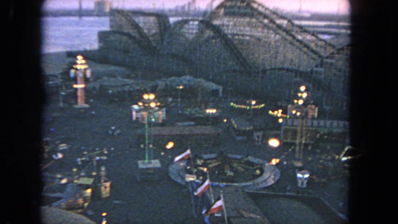 Amusement park seen from above with glowing lights, flags, and a wooden roller coaster behind the rides.