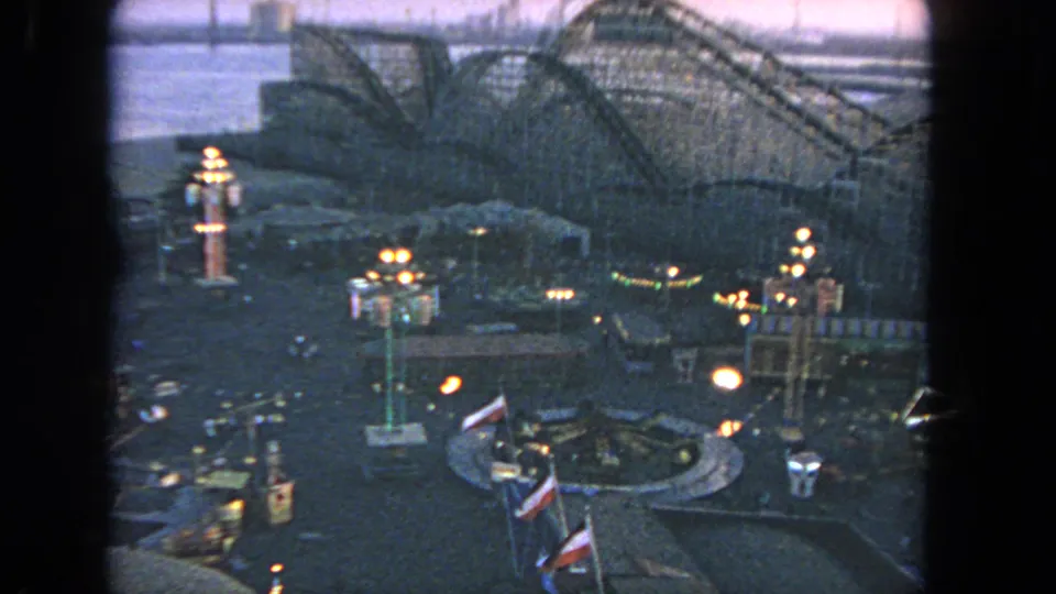 Elevated 1962 Los Angeles amusement-park view with lit midway structures, flags, and a wooden roller coaster silhouetted in dusk light.
