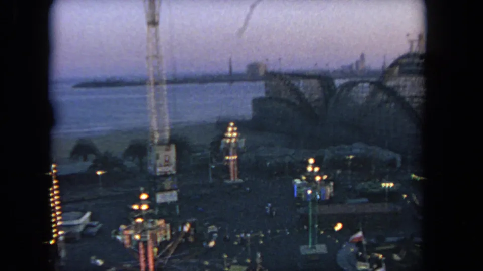 1962 Los Angeles amusement park scene with roller coaster framing, ride lights, water, and distant shoreline in low light.