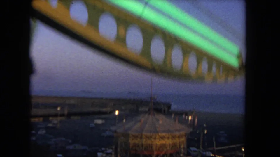 1962 coastal Los Angeles amusement view with Ferris wheel framing, carousel roof, pier, ocean, and evening lights.
